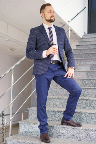 A young man in a business suit on the stairs with a glass of coffee. A businessman on the stairs.