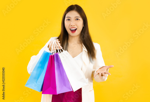 Young asian woman long hair carrying colorful paper shopping bags posing giving and presenting on yellow background.