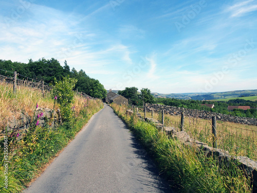 perspective view along a narrow country lane with a stone wall and farmhouse surrounded by fields in summer near the village of colden in calderdale west yorkshire