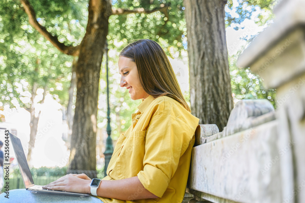 Happy female freelancer enjoying remote work sitting in the park, smiling woman using laptop computer outdoors, side view Happy female freelancer enjoying remote work sitting in the park, smiling woman using laptop computer outdoors, side view