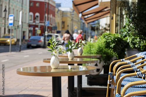 Street cafe in city with empty tables outdoor on walking people background. Vases of rose flowers on round tables and cozy chairs in sunny day © Oleg