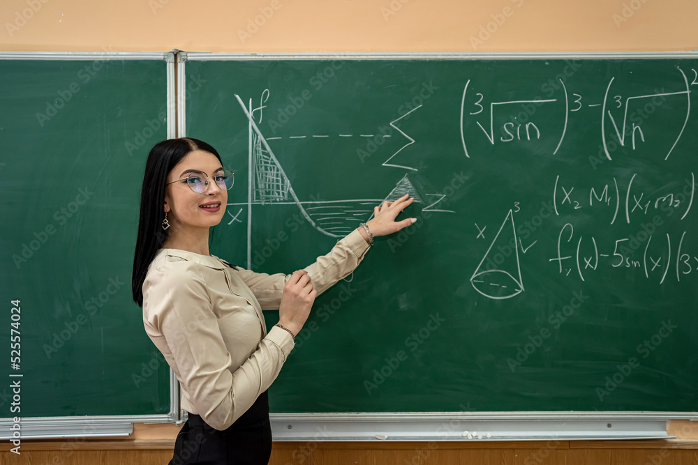 young pretty female teacher writing on a chalkboard completing mathematical equations in classroom young pretty female teacher writing on a chalkboard completing mathematical equations in classroom