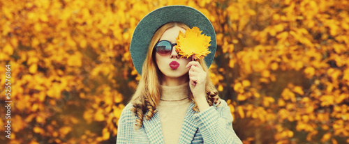 Autumn portrait of beautiful young woman with yellow maple leaves blowing her lips wearing round hat in the park