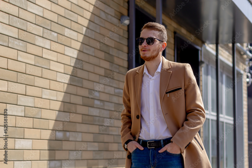 A young man dressed in a beige coat is walking down the street. A young bearded guy with a modern hairstyle and sunglasses on an urban background
