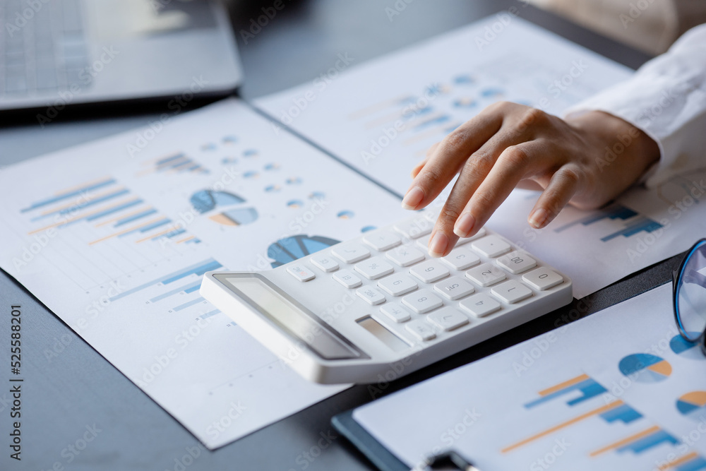 Businessman is using a calculator to calculate company financial figures from earnings papers, a businessman sitting in his office where the company financial chart is placed. Businessman is using a calculator to calculate company financial figures from earnings papers, a businessman sitting in his office where the company financial chart is placed.