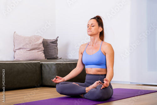 Fototapeta Naklejka Na Ścianę i Meble -  Calm woman dressed in sportswear, meditates on the floor in her living room, listening to spiritual practices classes, posing in lotus pose, tries to relax. Yoga concept