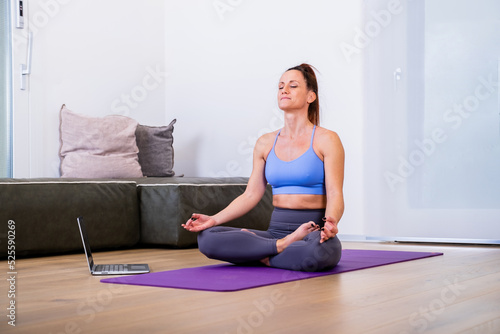 Calm woman dressed in sportswear, meditates on the floor in her living room, listening to spiritual practices classes on laptop, poses in lotus pose, tries to relax. Yoga concept