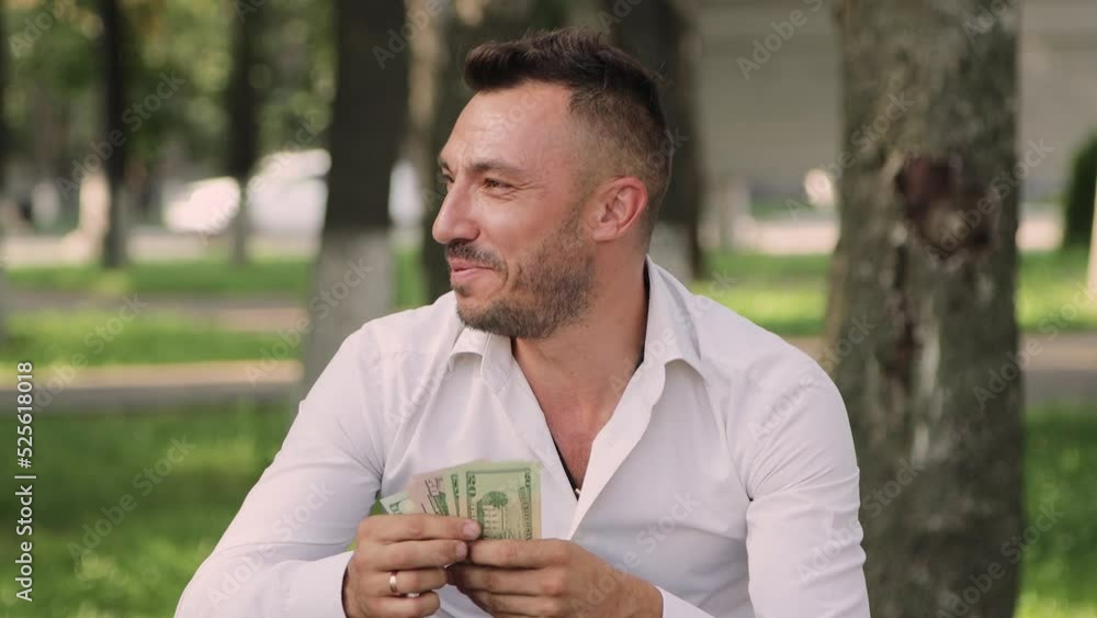 Close-up of a man laughing, counting dollars, he made a profit or a win. A man in a white shirt is sitting in a park on a bench against the background of green grass and trees. 4K UHD. Close-up of a man laughing, counting dollars, he made a profit or a win. A man in a white shirt is sitting in a park on a bench against the background of green grass and trees. 4K UHD.