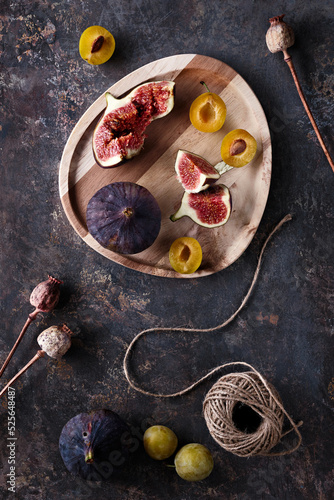 Autumn fruits, black fig and yellow plums on wooden tray. Flat lay, top view on dark textured background with dry poppy plants and hemp cord. Fruits in Fall season.