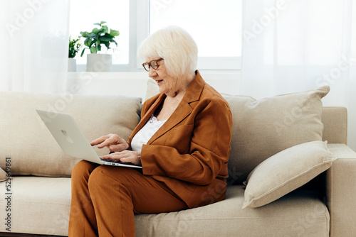 an elderly businesswoman is sitting on a beige sofa in her bright apartment and carefully looks at her laptop while working remotely behind it