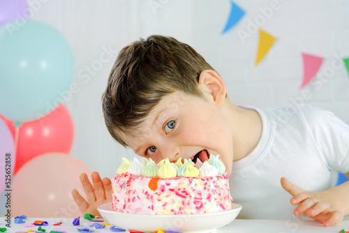 the boy is trying to take a bite out of the birthday cake. The child looks into the frame, against the background of balloons and multi-colored flags. Cheerful child indulges in his birthday.