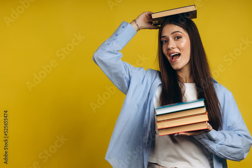 Portrait of student girl balancing books on head and hand while standing against yellow background