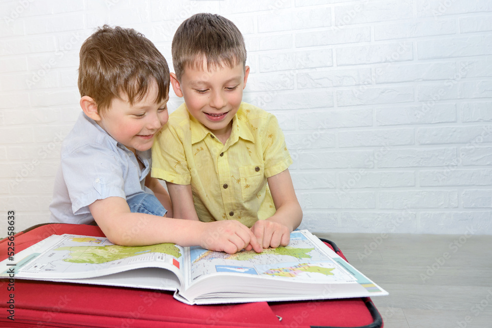 two cheerful brothers sit on a suitcase and look at an atlas, children dream of traveling. The boys laugh and smile. Horizontal photo. two cheerful brothers sit on a suitcase and look at an atlas, children dream of traveling. The boys laugh and smile. Horizontal photo.