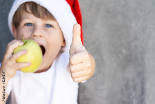 A child with a green apple in a Santa Claus hat shows a thumbs up to the dentist and healthy white teeth.