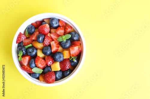 Fruit salad in a white cup on a yellow background. Sliced strawberries  peaches and blueberries with mint leaves. Free space for text.