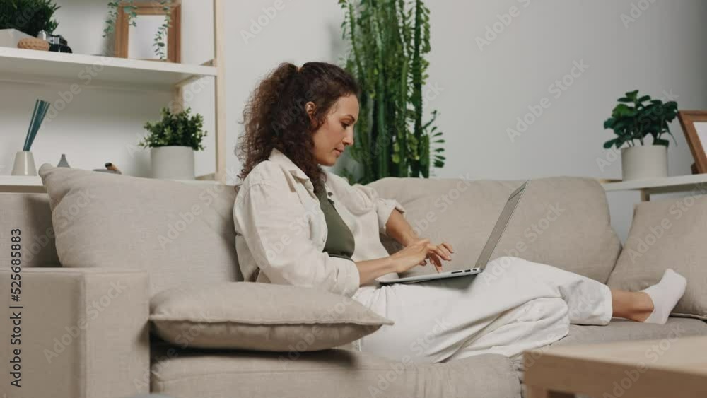 Freelance woman working on a conference project and typing on a gray laptop on a keyboard with her laptop on her lap at home on the couch at night Freelance woman working on a conference project and typing on a gray laptop on a keyboard with her laptop on her lap at home on the couch at night