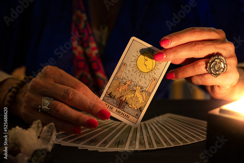 Fortune teller hands showing The Moon tarot card, symbol of intuition, during a reading. Close-up with candle light, moody atmosphere.