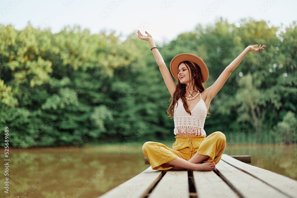 Hippie eco-activist woman traveler sits on a bridge by a lake with her arms outstretched with a hat and smiling sincerely