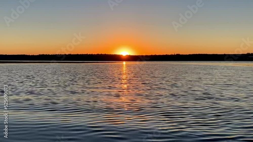 Sunrise over the river near the shore. Rising sun in clear sky. Changeable weather, calm water and waves on the lake. Reflection and glare in the sunlight. Timelapse, 4k.