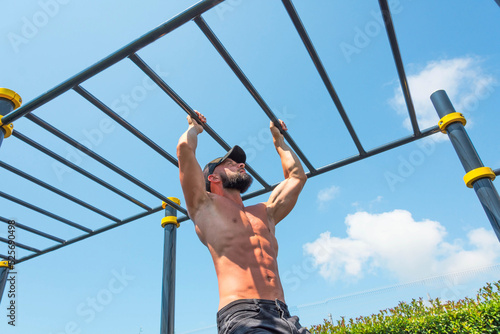 Muscular man in a cap hanging on the stairs intercepted on the rungs with his hands in the park outdoors.