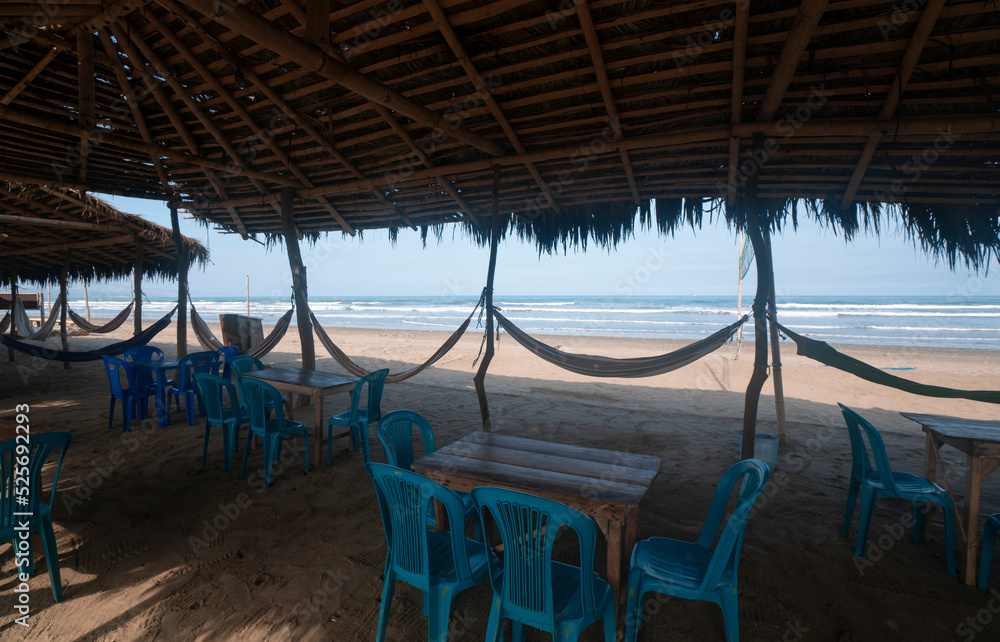 View of a rustic restaurant without people, with a bamboo roof, wooden tables, plastic chairs and hammocks hanging from poles by the beach
