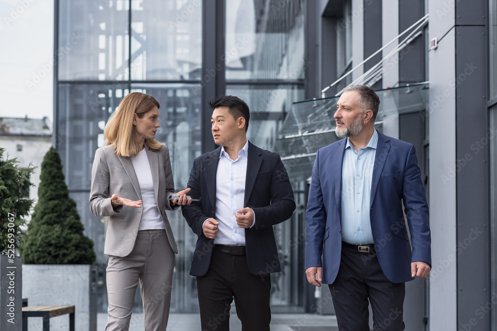 Three diverse business people walking and talking focused and thoughtful seriously outside office building, male and female, discussing plans and work project Three diverse business people walking and talking focused and thoughtful seriously outside office building, male and female, discussing plans and work project