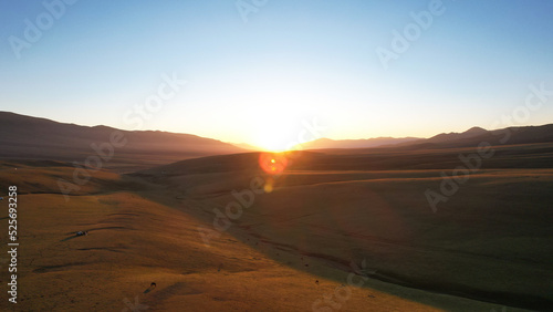Bright yellow dawn with a red tint in the mountains. The green hills with grass turned red. A small stream flows through the gorge  horses graze. Clear blue sky. View from top. Assy Plateau Kazakhstan