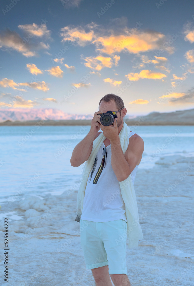 Obraz premium Adult man photographer taking photographs with digital camera at Dead Sea beach in Ein Bokek, Israel. Handsome muscular man using picture with modern dslr camera at the beach of seashore. Travel