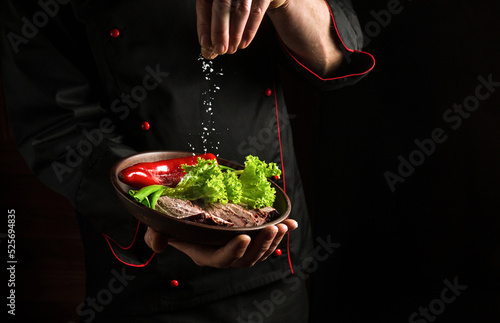 Professional chef sprinkles salt on a sliced steak with beef and vegetables in a plate. The concept of serving dishes to order
