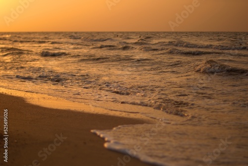 The sandy shore of the Sea of Azov at sunset, a warm summer evening, the waves are beating against the shore, stroking the sand with sea foam, defocus, selective focus.