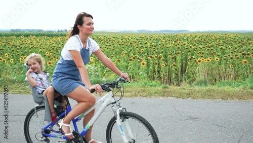 Slow motion of mother riding bicycle and little girl sitting in child seat behind her against the backdrop of a blooming sunflower field. Tracking shot of happy family enjoying bike ride Outdoors.