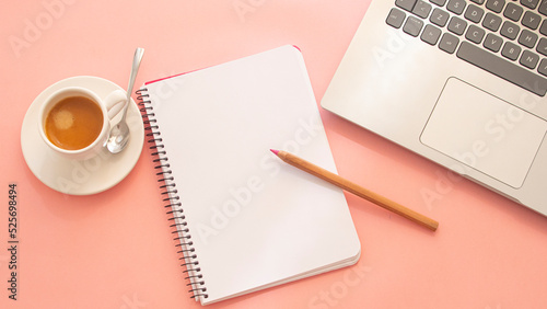 Blank page of a notebook next to a laptop and a white cup with coffee on a light pink background