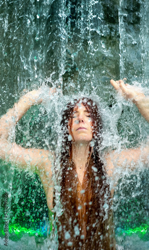 beautiful young sexy redhead woman under the splashing water of the waterfall  with green light  in the Spa Wellness pool  raises her arms  closes her eyes under the rippling water