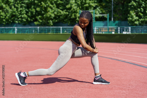 African American lady in sportswear pumps gluteal muscles on sports ground near lush green trees growing in city park. Black woman with long braids enjoys effective exercise on sunny summer day