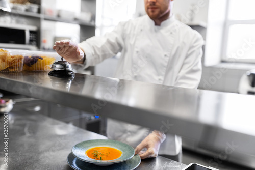 food cooking, profession and people concept - close up of male chef with plate of pumpkin cream soup ringing bell at restaurant kitchen table