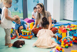 Toddlers and their nursery teacher playing with plastic building blocks, colorful cars and other toys while sitting on the floor in a play room. Fine motor and gross motor skills development, problem