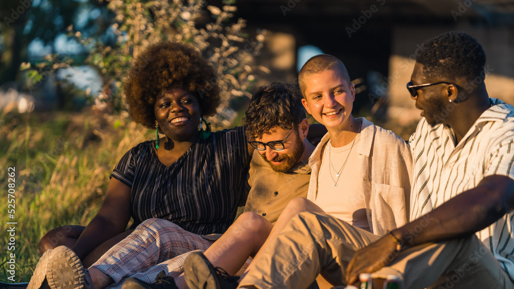 Obraz premium Group of happy young people having a weekend picnic in nature, sitting together on the grass near the campfire, talking and laughing. Sunset time. High quality photo