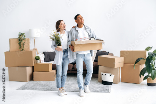 Happy multiracial young couple in love walk into their new home, holding boxes of household items, look around the interior with excited, rejoice at their new home, smile. Moving to a new apartment © Kateryna