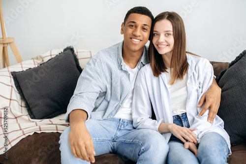 Happy together. Portrait of married young couple. Positive multiracial couple in love, caucasian woman and hispanic man, sitting on sofa at home in living room, hugging, looking at camera, smiling 