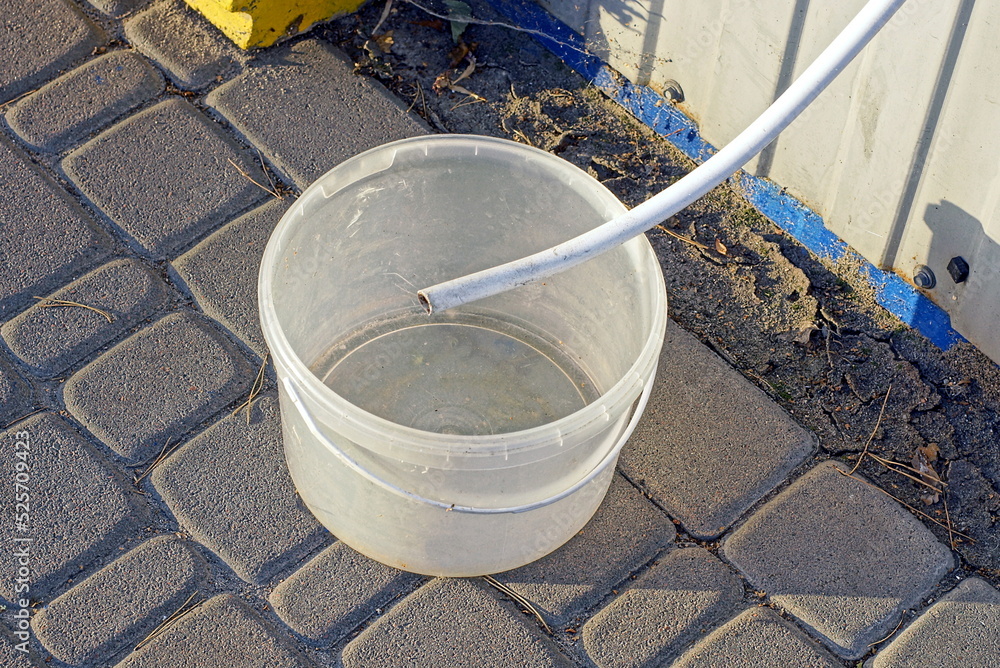 a white water hose in an empty plastic bucket stands on a gray sidewalk in the street