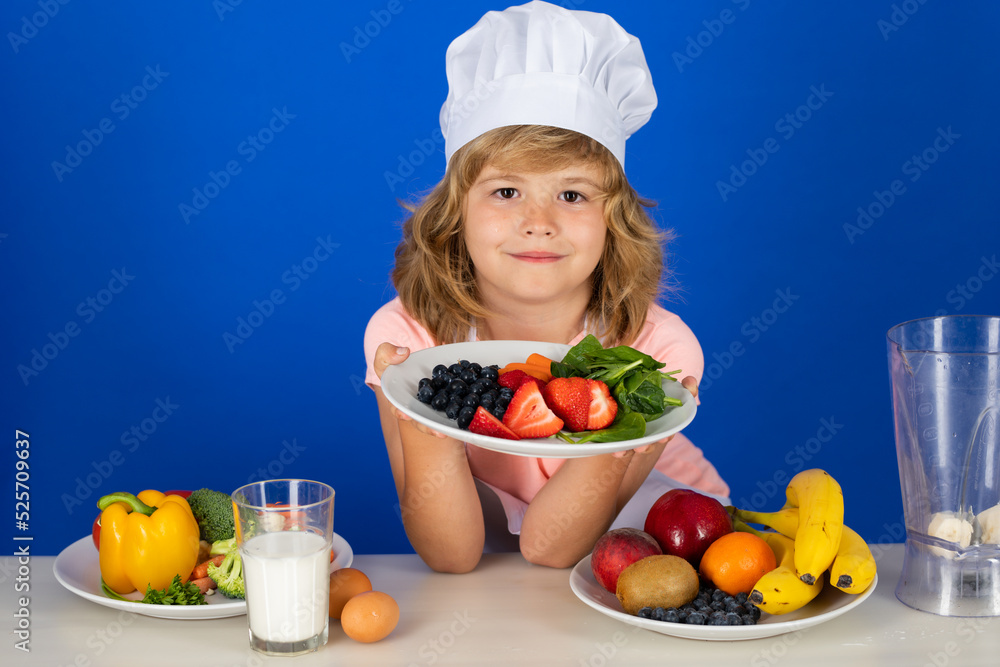 Child chef cook hold plate with fruits prepares food in isolated blue studio background. Kids cooking. Teen boy with apron and chef hat preparing a healthy vegetables meal in the kitchen. Child chef cook hold plate with fruits prepares food in isolated blue studio background. Kids cooking. Teen boy with apron and chef hat preparing a healthy vegetables meal in the kitchen.