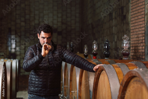 Man tasting red wine in a winery barrel cellar, holding a wine glass, swirling it, smelling, and sipping wine, delighted with wine taste and flavor.