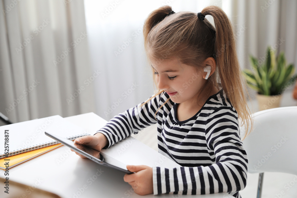 Adorable little girl doing homework with tablet and earphones at table indoors Adorable little girl doing homework with tablet and earphones at table indoors