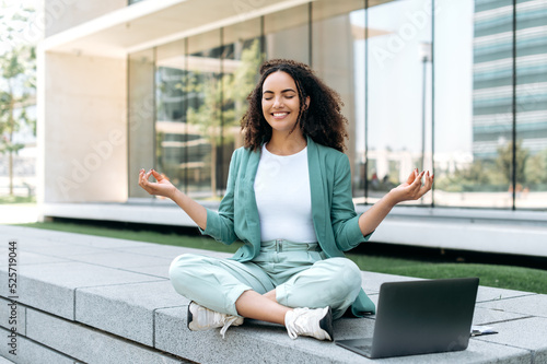 Break during work. Calm relaxed curly haired hispanic or brazilian young woman, office worker, sits in a lotus position outdoors near the business center, meditates, relaxes, closed her eyes, smile Free Download Break during work. Calm relaxed curly haired hispanic or brazilian young woman, office worker, sits in a lotus position outdoors near the business center, meditates, relaxes, closed her eyes, smile - GFXLoad