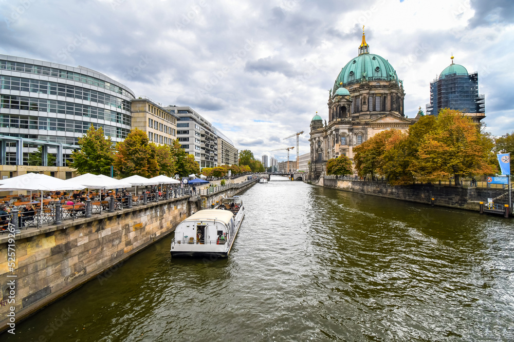 A tour boat alongside cafe patios on the Spree River at Autumn with the Cathedral in view in Berlin, Germany.