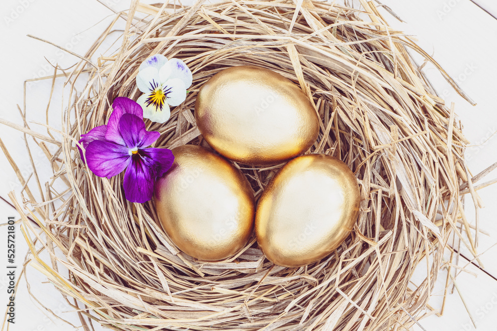 Obraz premium Easter golden eggs in a hay nest with flowers, overhead view on white background