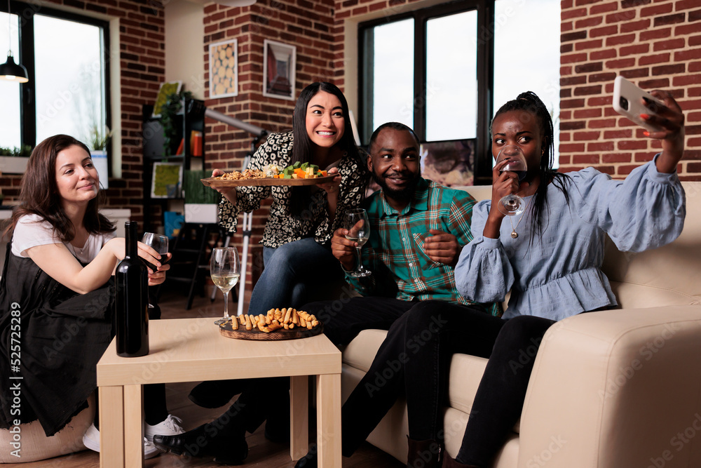 Multiracial cheerful young people at home celebrating birthday while taking selfie photos together and drinking wine. Group of friends drinking alcoholic beverages while photographing themselves. Multiracial cheerful young people at home celebrating birthday while taking selfie photos together and drinking wine. Group of friends drinking alcoholic beverages while photographing themselves.