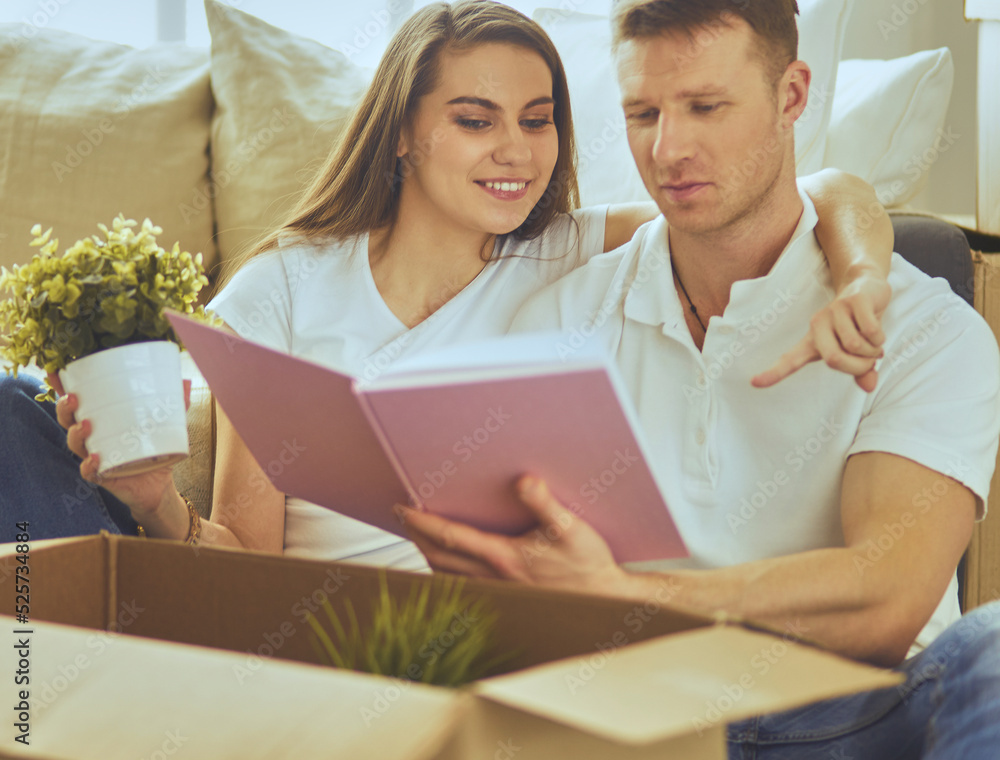 Cute couple unpacking cardboard boxes in their new home, sitting on the floor and looking at a family album Cute couple unpacking cardboard boxes in their new home, sitting on the floor and looking at a family album