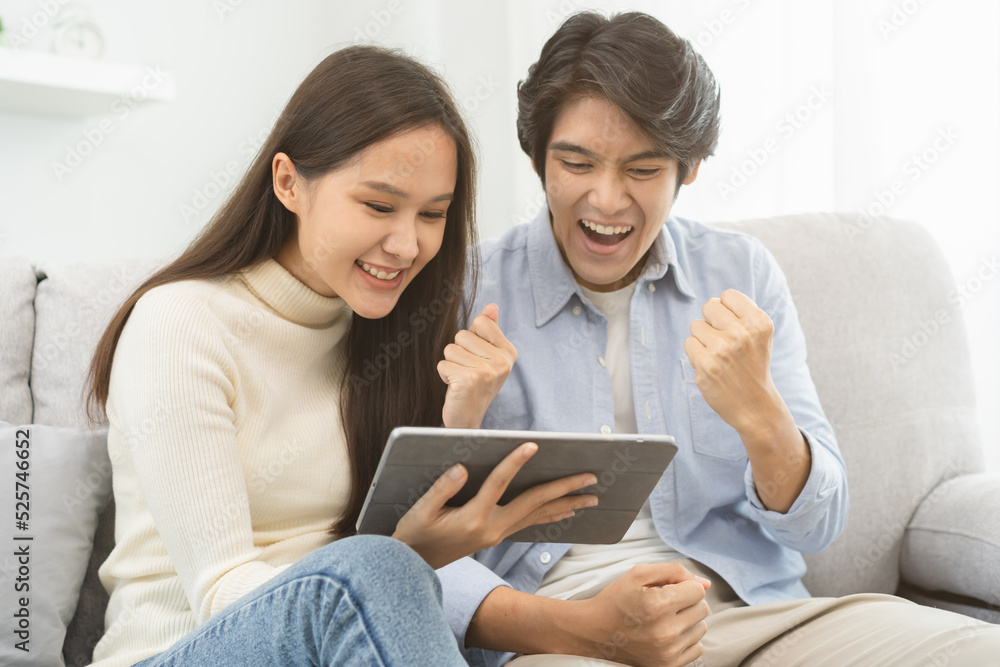 Happy excited, smiling asian young couple love using tablet pc, great deal or business success, received or getting cash back, tax refund, good news by mail while sitting on sofa, couch at home.