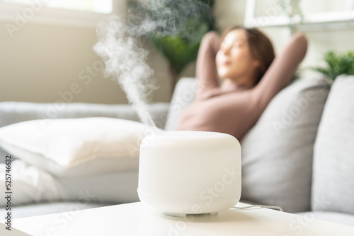 Modern air humidifier during relax or rest, happy blurred asian young woman, girl enjoying aromatherapy steam scent from essential oil diffuser comfortable in living conditions room,apartment at home photo